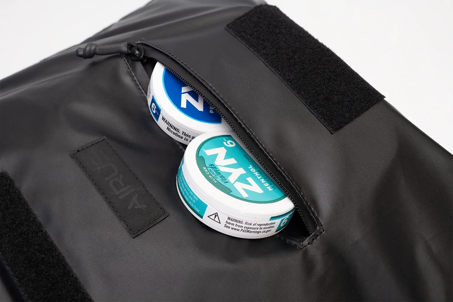 The zipped front pocket of the AIRUS carry bag, half unzipped, with two cans of nicotine pouches inside.
