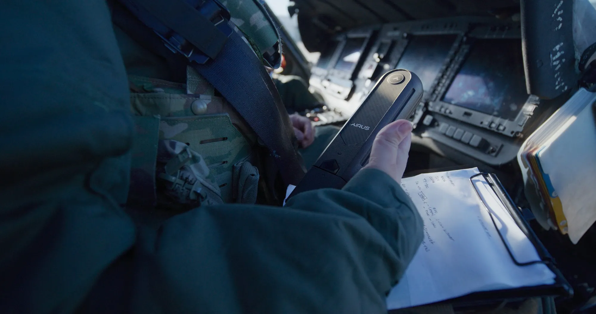 An aircrew member holds the AIRUS controller in a helicopter cockpit, a kneeboard open beside them.