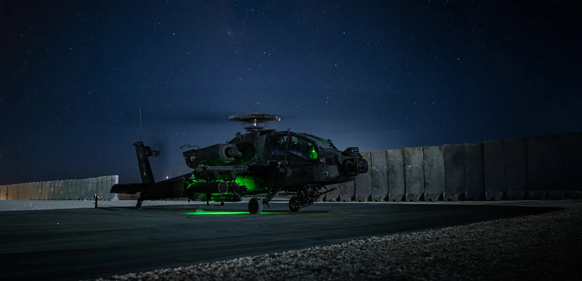 An AH-64 Apache on a landing pad at night, lit from beneath, under a clear sky.