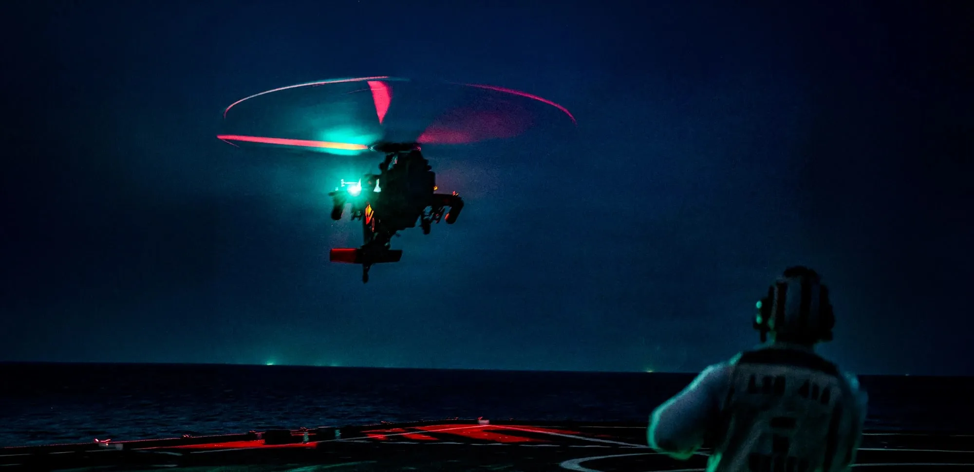 A helicopter on approach to a ship’s flight deck at night, guided in by a deck crewman under low-visible lighting.