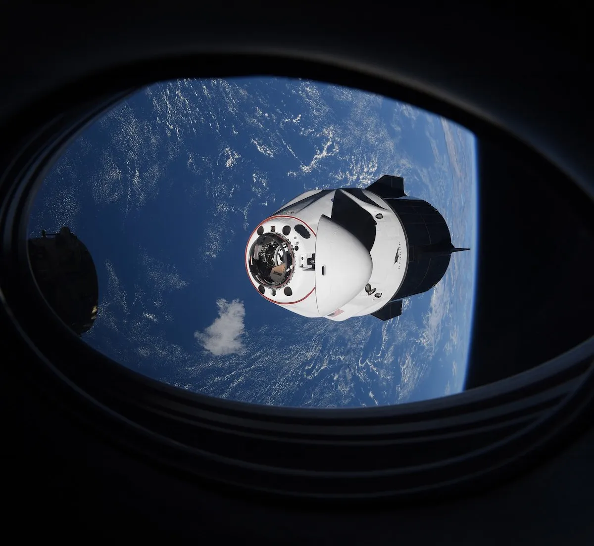 A SpaceX Crew Dragon spacecraft in orbit above Earth, seen through a window of the International Space Station.