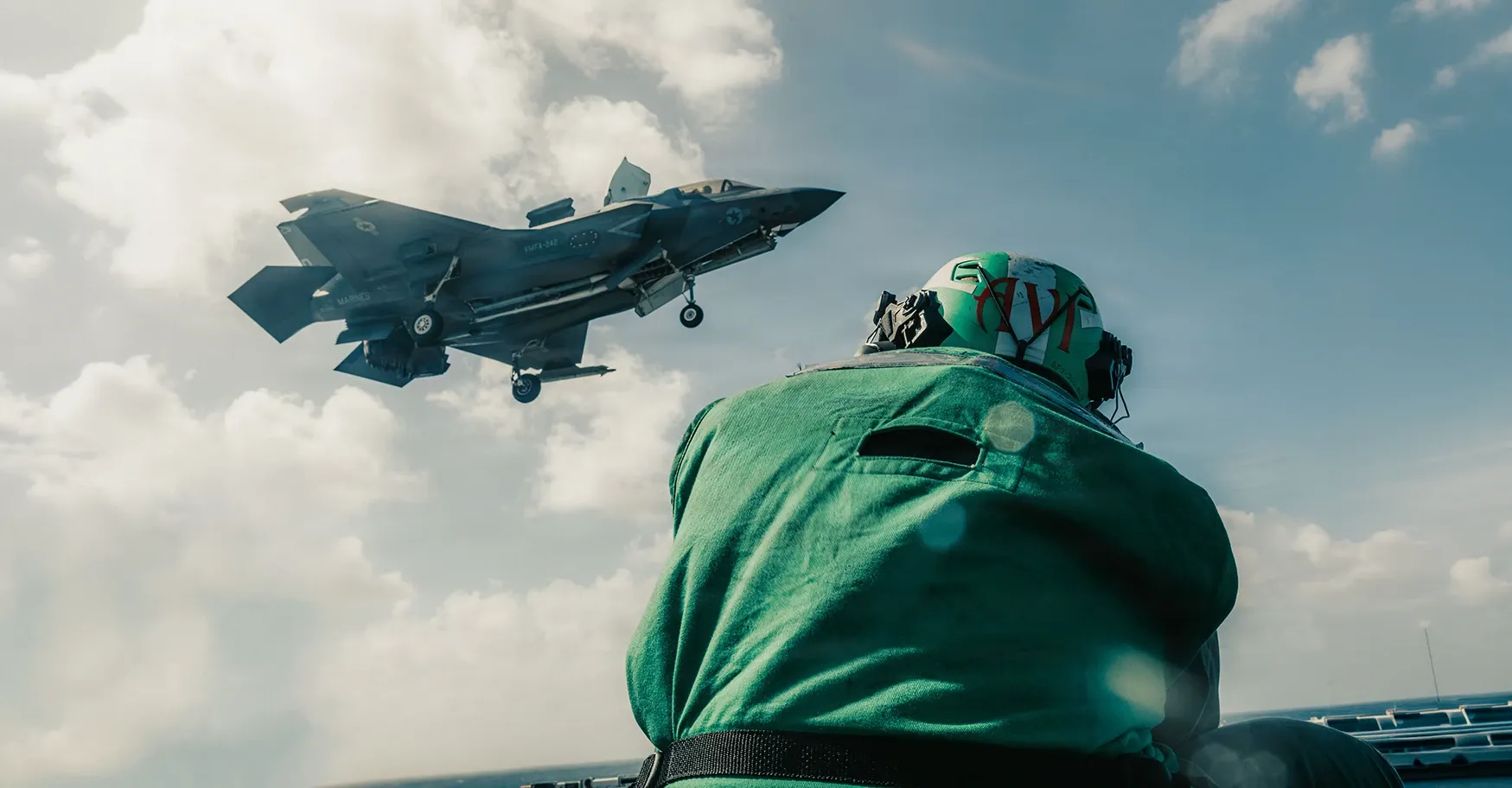 A U.S. Marine Corps F-35B, lift fan open and gear down, over a ship’s flight deck past a crew member in a green deck jersey.